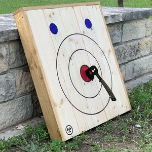May include: Axe throwing target made of light-colored wood, featuring concentric circles and a red bullseye. Two blue circles are at the top. An axe is embedded in the center. The target leans against a stone wall.