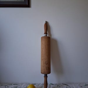 Può includere: Un mattarello in legno con manici marrone scuro e una superficie di rotolamento cilindrica. Il mattarello è in posizione verticale contro un muro bianco. Un limone giallo è appoggiato su una tovaglia bianca con motivi floreali ricamati.