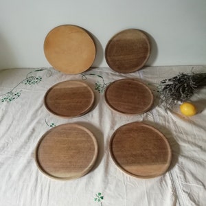 Op de afbeelding: Zes ronde houten serveerschalen, vijf zijn donkerbruin en één is lichtbruin. De schalen zijn gerangschikt op een wit tafelkleed met groene bloemenborduursels.