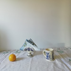 May include: A white ceramic napkin holder with blue floral designs, a white ceramic mug with blue floral designs, and a yellow lemon on a white tablecloth with a green floral pattern.