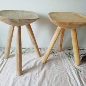 May include: Two wooden stools with a bowl-shaped seat and three legs. The stools are made of natural wood and have a rustic look.