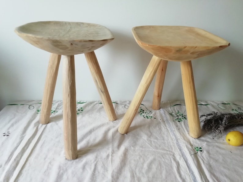 May include: Two wooden stools with a bowl-shaped seat and three legs. The stools are made of natural wood and have a rustic look.