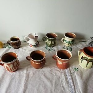 以下が含まれることがあります： さまざまなデザインの手描きセラミックマグカップとピッチャー8個のセット。マグカップとピッチャーは、花柄と幾何学模様が施された茶色、白、緑色です。