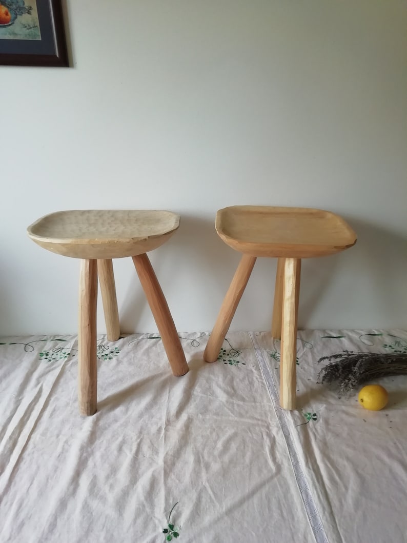 May include: Two wooden stools with a natural finish and three legs each. The stools are sitting on a white tablecloth with a floral pattern.
