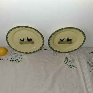 Puede incluir: Dos platos ovalados con superficie crema y borde festoneado verde. Cada plato presenta una escena pintada de dos gallinas sobre un césped. Un limón descansa sobre un mantel blanco.
