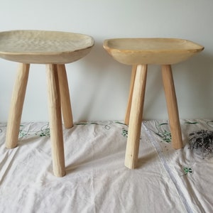 May include: Two wooden stools with a unique, carved bowl-shaped seat. The stools have three legs each and are made of natural wood.