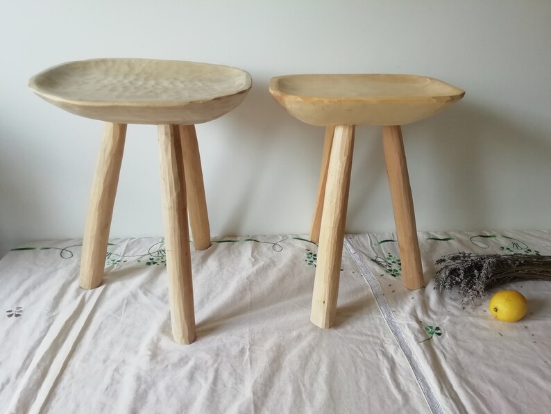 May include: Two wooden stools with a unique, carved bowl-shaped seat. The stools have three legs each and are made of natural wood.