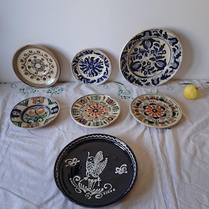 Può includere: Una collezione di sette piatti rotondi in ceramica con vari disegni dipinti a mano. I piatti presentano motivi complessi in blu, verde, arancione e marrone, inclusi disegni floreali, forme geometriche e l'illustrazione di un uccello su un piatto blu scuro.