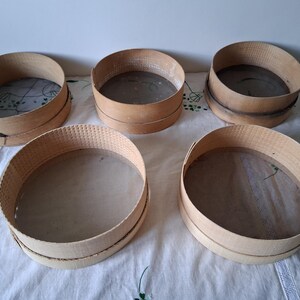May include: Five wooden flour sifters with mesh bottoms. The sifters are round and have a natural wood finish. They are arranged in a circle on a white tablecloth.