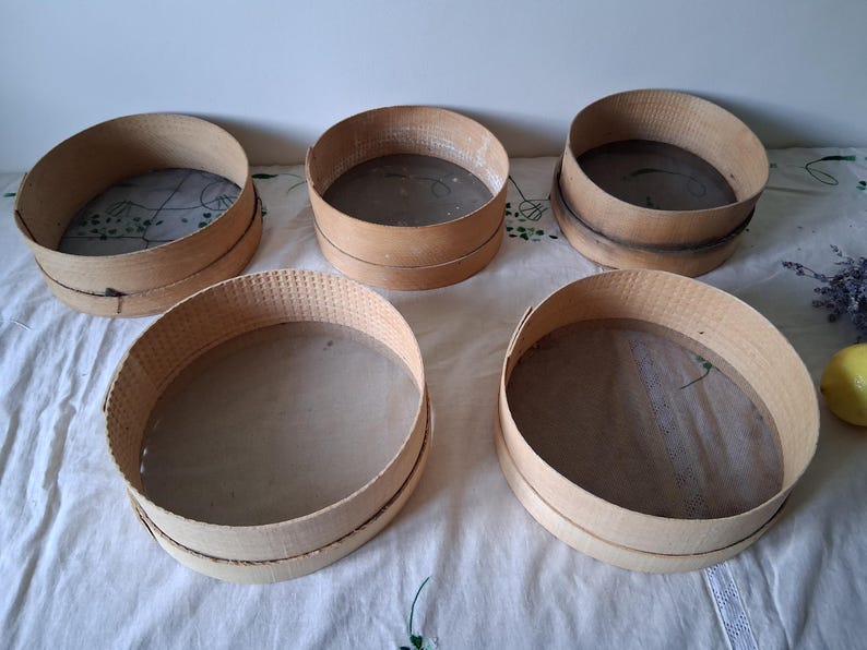 May include: Five wooden flour sifters with mesh bottoms. The sifters are round and have a natural wood finish. They are arranged in a circle on a white tablecloth.