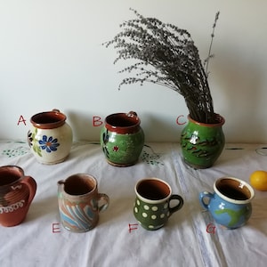Può includere: Sette tazze in ceramica fatte a mano con vari disegni e colori. Le tazze sono disposte su una tovaglia bianca con un mazzo di lavanda secca in un vaso di ceramica verde.