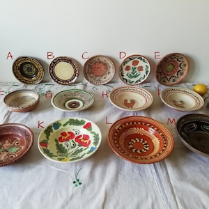 May include: A collection of 13 hand-painted ceramic bowls in various colors and designs. The bowls are arranged in two rows on a white tablecloth with a floral pattern. Some of the bowls feature floral designs, while others have geometric patterns or animal motifs.