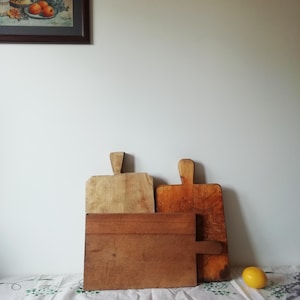 Può includere: Tre taglieri di legno, uno marrone, uno marrone chiaro e uno con finitura in legno naturale, sono impilati su una tovaglia bianca. Un limone giallo si trova sulla tovaglia.