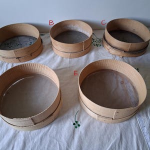 May include: Five wooden flour sifters with mesh screens. The sifters are stacked on a white tablecloth.