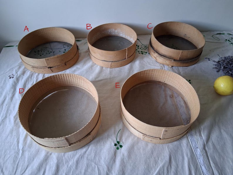 May include: Five wooden flour sifters with mesh screens. The sifters are stacked on a white tablecloth.