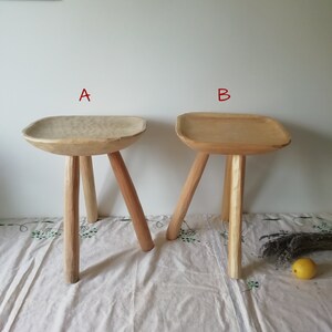 May include: Two wooden stools with three legs each. The stool on the left has a rounded top, while the stool on the right has a square top. Both stools are made of light-colored wood.