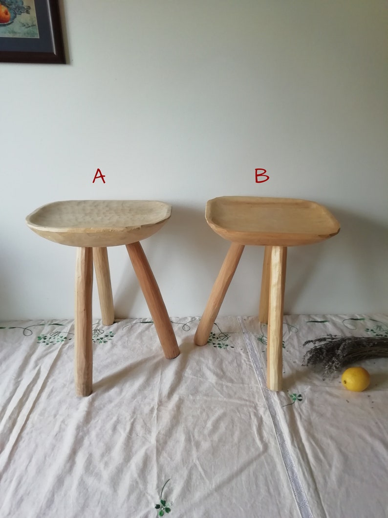 May include: Two wooden stools with three legs each. The stool on the left has a rounded top, while the stool on the right has a square top. Both stools are made of light-colored wood.