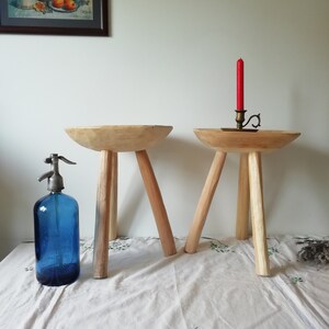 May include: Two wooden stools with three legs each. The stools are made of natural wood and have a rustic look. One stool has a red candle on top of it.