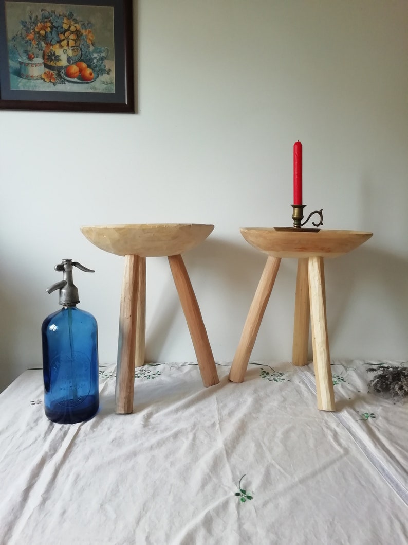 May include: Two wooden stools with three legs each. The stools are made of natural wood and have a rustic look. One stool has a red candle on top of it.