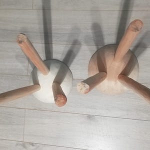 May include: Two wooden stools with three legs each. The stools are made of light wood and have a white, round seat. The stools are sitting on a light wood floor.