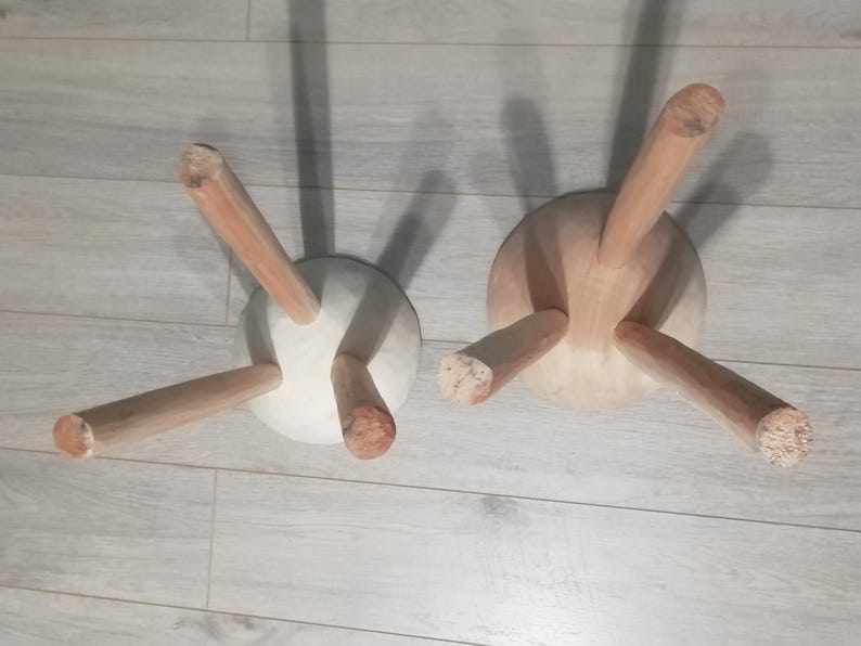 May include: Two wooden stools with three legs each. The stools are made of light wood and have a white, round seat. The stools are sitting on a light wood floor.