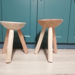 May include: Two wooden stools with three legs each. The stools are made of natural wood and have a light brown color. The stools are sitting on a light brown floor.