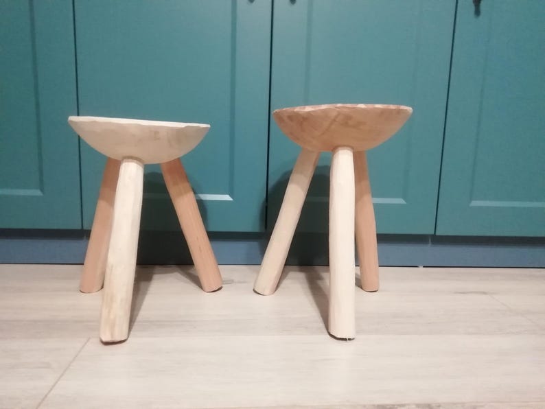 May include: Two wooden stools with three legs each. The stools are made of natural wood and have a light brown color. The stools are sitting on a light brown floor.
