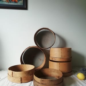 Może przedstawiać: Zestaw sześciu drewnianych sit do mąki w r&oacute;żnych rozmiarach. Sita wykonane są z naturalnego drewna i mają rustykalny, vintage'owy wygląd. Są ułożone jedno na drugim, z największym sitkiem na dole i najmniejszym na g&oacute;rze.
