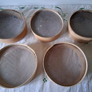 May include: Five round wooden sifters with mesh bottoms. The sifters are all the same size and are arranged in a semi-circle on a white tablecloth.
