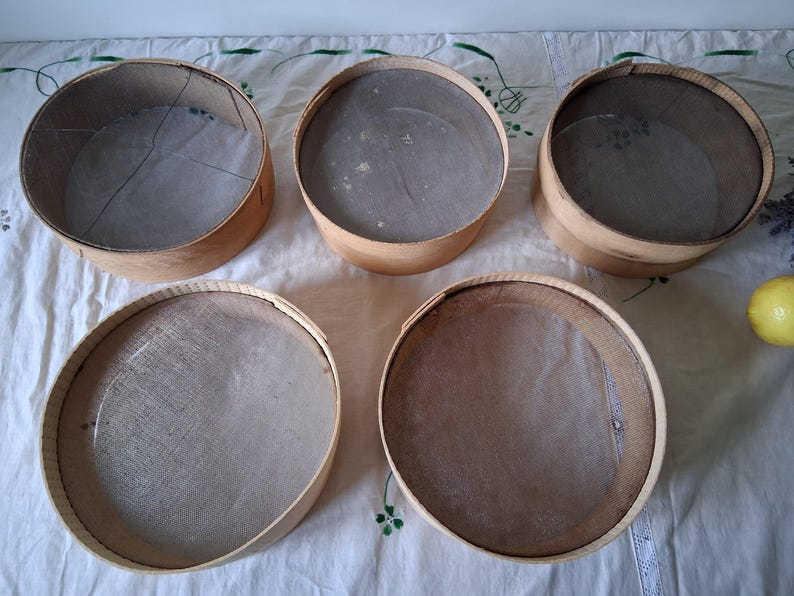 May include: Five round wooden sifters with mesh bottoms. The sifters are all the same size and are arranged in a semi-circle on a white tablecloth.