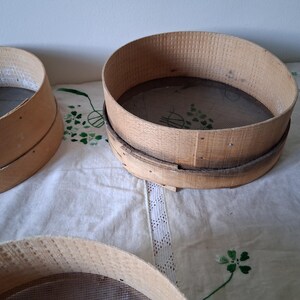 May include: Three wooden flour sifters with mesh screens. The sifters are round and have a natural wood finish. They are sitting on a white tablecloth with a lace trim.