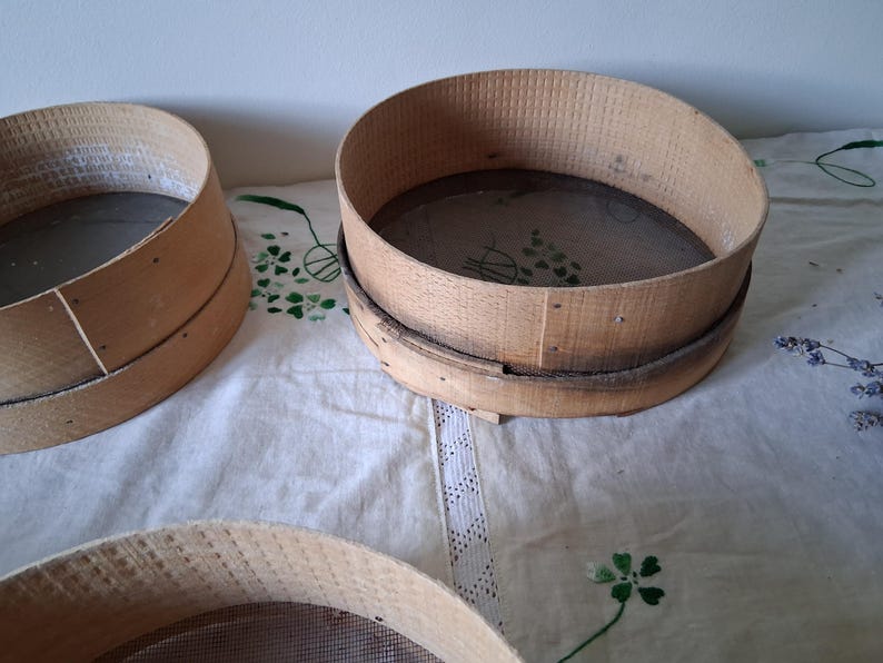May include: Three wooden flour sifters with mesh screens. The sifters are round and have a natural wood finish. They are sitting on a white tablecloth with a lace trim.