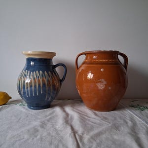 Può includere: Due vasi in ceramica, uno blu con disegni bianchi e arancioni e uno marrone con un bordo bianco. I vasi sono su una tovaglia bianca.