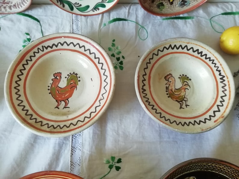 May include: Two white ceramic bowls with orange and green rooster designs. The bowls have a brown and black wavy border.