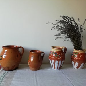 May include: Four brown pottery jugs with painted designs. The jugs are arranged in a row on a white tablecloth. A bunch of dried lavender is in the background.