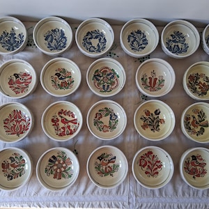 Può includere: Una collezione di ventuno piccole ciotole rotonde in ceramica con disegni floreali e di uccelli dipinti a mano. Le ciotole presentano una base color crema con motivi colorati in blu, rosso, verde e giallo. Le ciotole sono disposte su una tovaglia bianca.