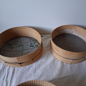 May include: Two round wooden flour sifters with mesh screens. The sifters are made of natural wood and have a rustic appearance.