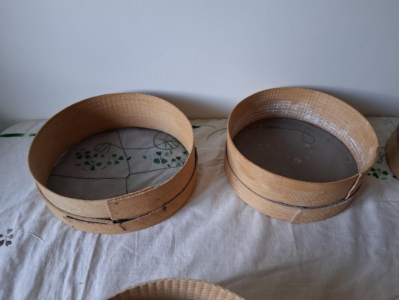 May include: Two round wooden flour sifters with mesh screens. The sifters are made of natural wood and have a rustic appearance.