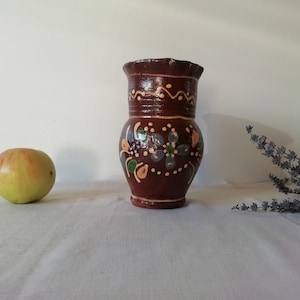 Könnte beinhalten: Ein brauner Keramik Krug mit einem Blumendesign in Blautönen, Grün und Gelb. Der Krug hat einen leicht ausgestellten Rand und einen runden Körper.
