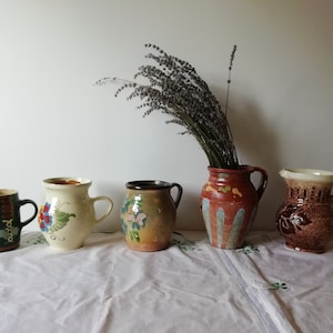 Può includere: Cinque brocche in ceramica con diversi disegni e colori. Le brocche sono disposte in fila su una tovaglia bianca. Un mazzo di lavanda secca è nel vaso a destra.