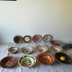 May include: A collection of 13 hand-painted ceramic bowls with various designs, including floral, geometric, and rooster motifs. The bowls are in shades of brown, red, green, blue, and white.