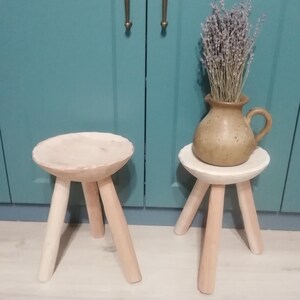 May include: Two wooden stools with three legs each. The stools are painted a light wood color and have a round top. The stools are sitting on a light wood floor. A vase with dried lavender is sitting on the top of one of the stools.