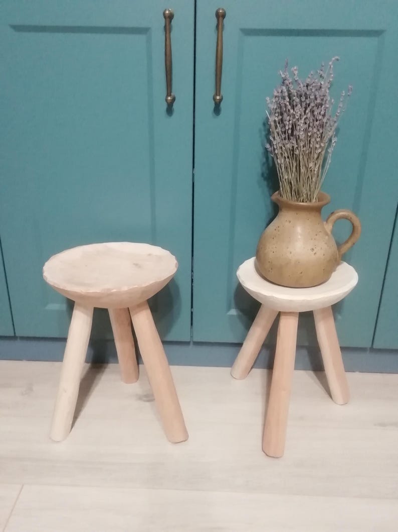May include: Two wooden stools with three legs each. The stools are painted a light wood color and have a round top. The stools are sitting on a light wood floor. A vase with dried lavender is sitting on the top of one of the stools.