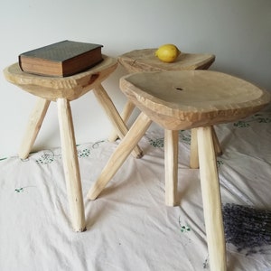May include: Three wooden stools, each with three legs. The stools are made from natural wood and have a rustic, handmade appearance. The stools are arranged in a row on a white tablecloth with a lemon on top of one of the stools.