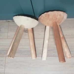 May include: Two wooden stools with three legs each. One stool has a white top and the other has a natural wood top.