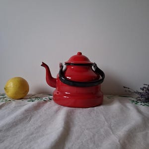 Può includere: Una teiera di smalto rosso con manico e coperchio neri. La teiera è appoggiata su una tovaglia bianca con un limone accanto.