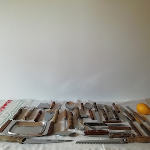 Può includere: Un set di utensili da cucina vintage con manici in legno, tra cui coltelli, forchette, cucchiai e spatole. Gli utensili sono disposti su una tovaglia bianca con un motivo rosso e verde.