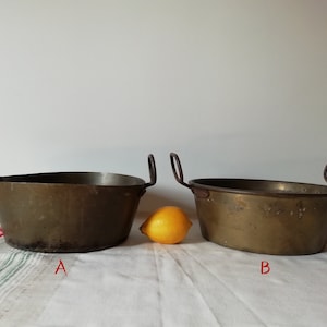 Puede incluir: Dos ollas de cobre antiguas con asas de hierro. Las ollas son de diferentes tamaños y tienen un aspecto desgastado y envejecido. La olla más grande está etiquetada con "B" y la olla más pequeña está etiquetada con "A".