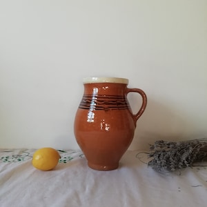 May include: A brown ceramic pitcher with a white rim and brown stripes around the body. The pitcher has a handle and is sitting on a white tablecloth.