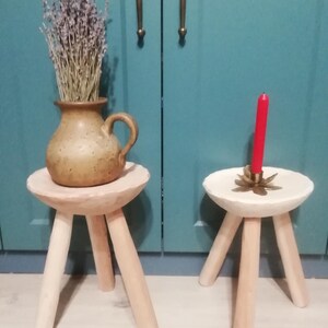 May include: Two wooden stools with three legs each. The stool on the left has a light brown top and a brown ceramic vase with dried lavender on top. The stool on the right has a white top and a red candle on top.